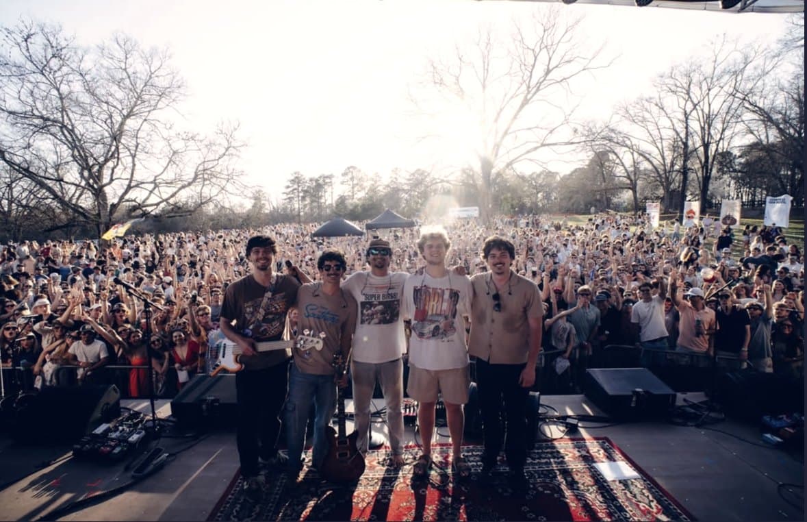 A band standing shoulder to shoulder at the front of the stage as the crowd stretches back into the trees.