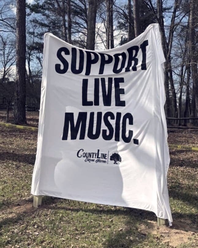 A canvas banner reading Support Live Music, hung between two pines at the edge of the field.
