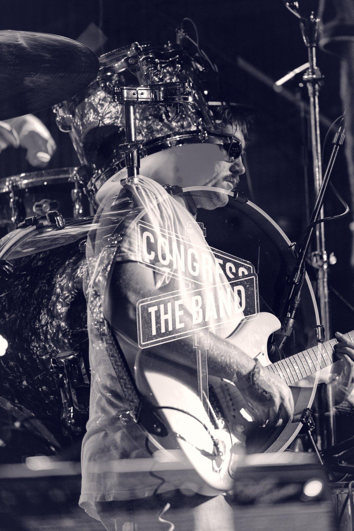 A guitarist photographed in black and white against the dark of an indoor stage.