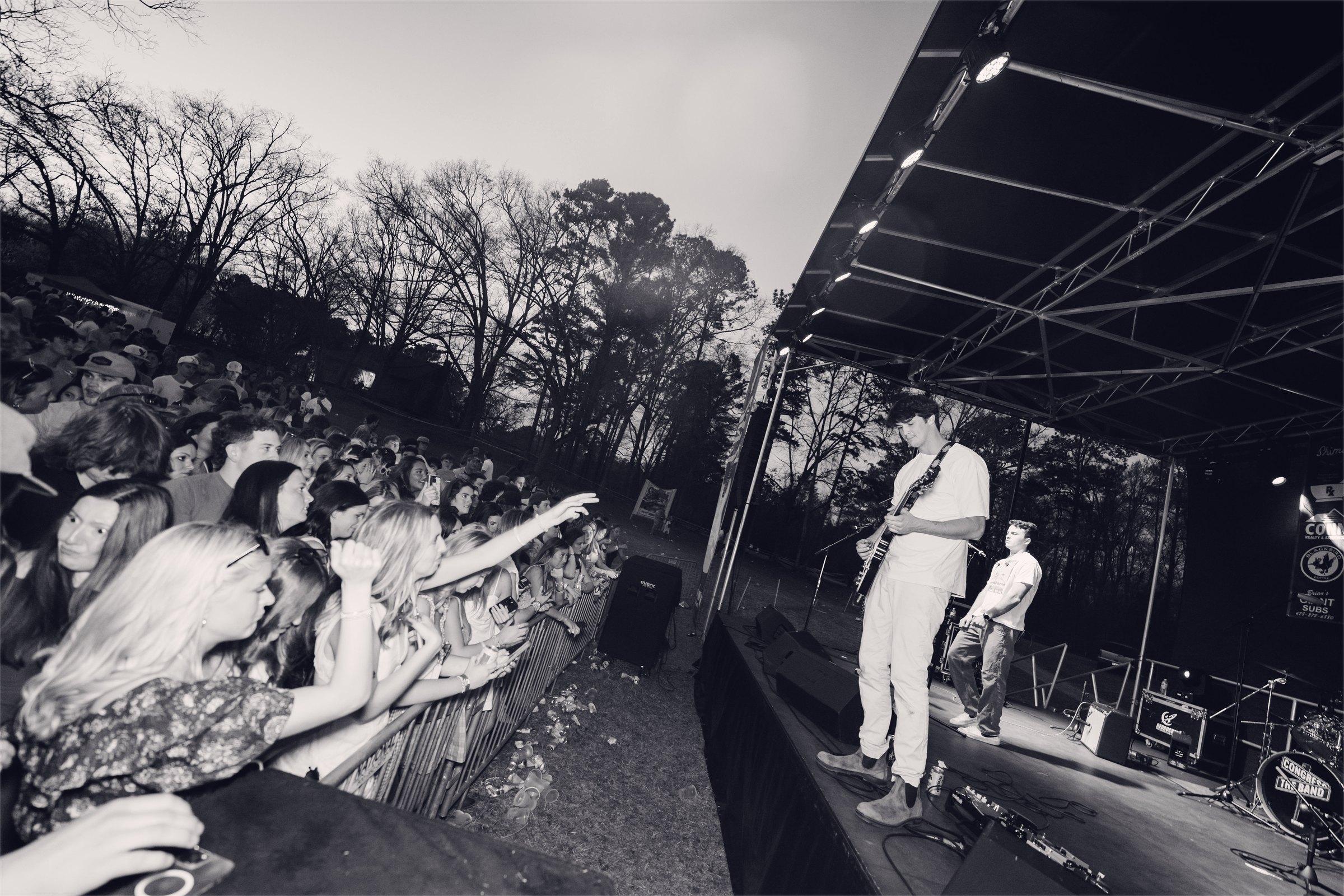 A black-and-white view from the side of the stage, the crowd running back through bare pines.