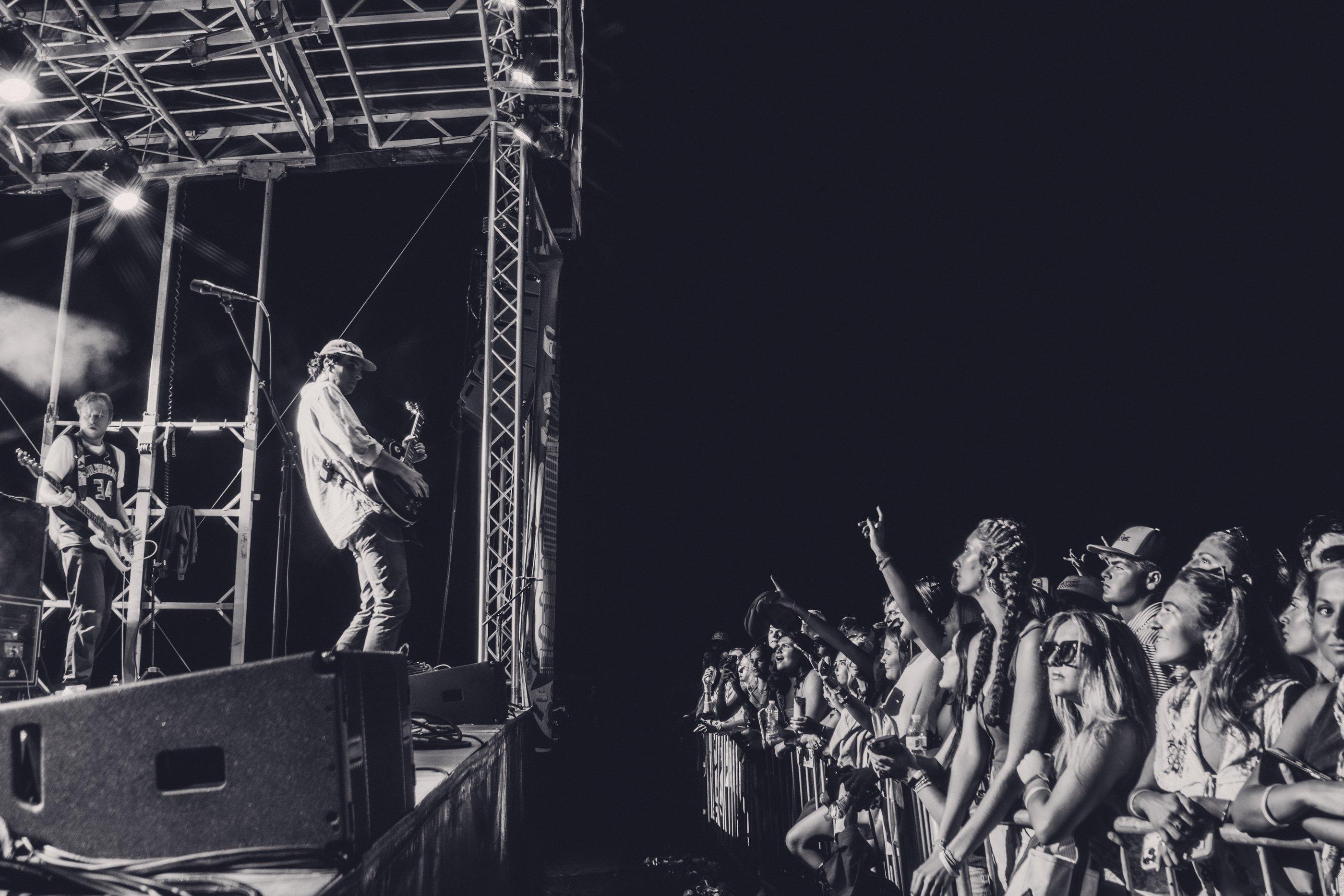 A guitarist lit at the edge of a night stage while the front row watches from the barricade.