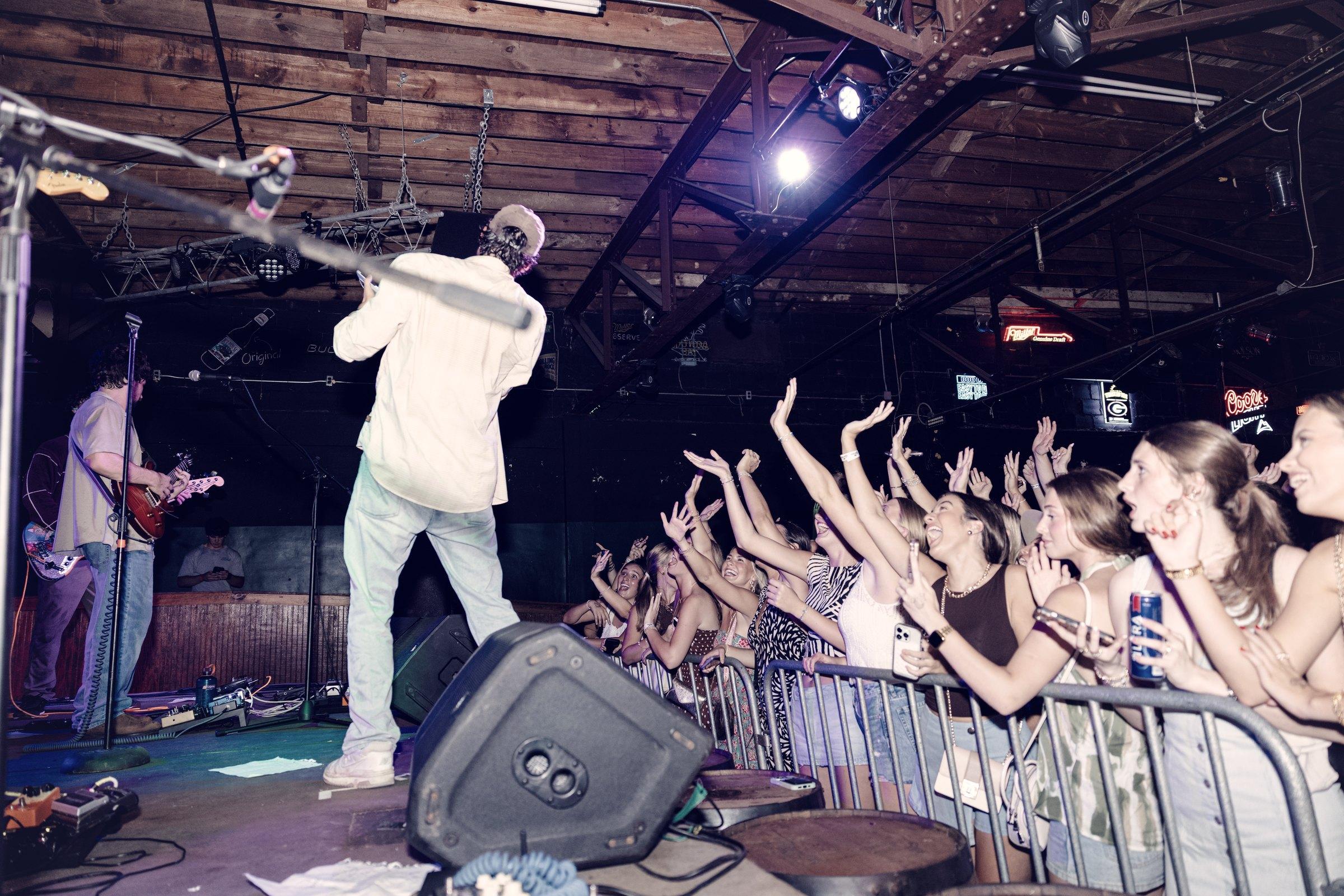 A performer leaning out over the barricade toward a packed room under exposed timber trusses.