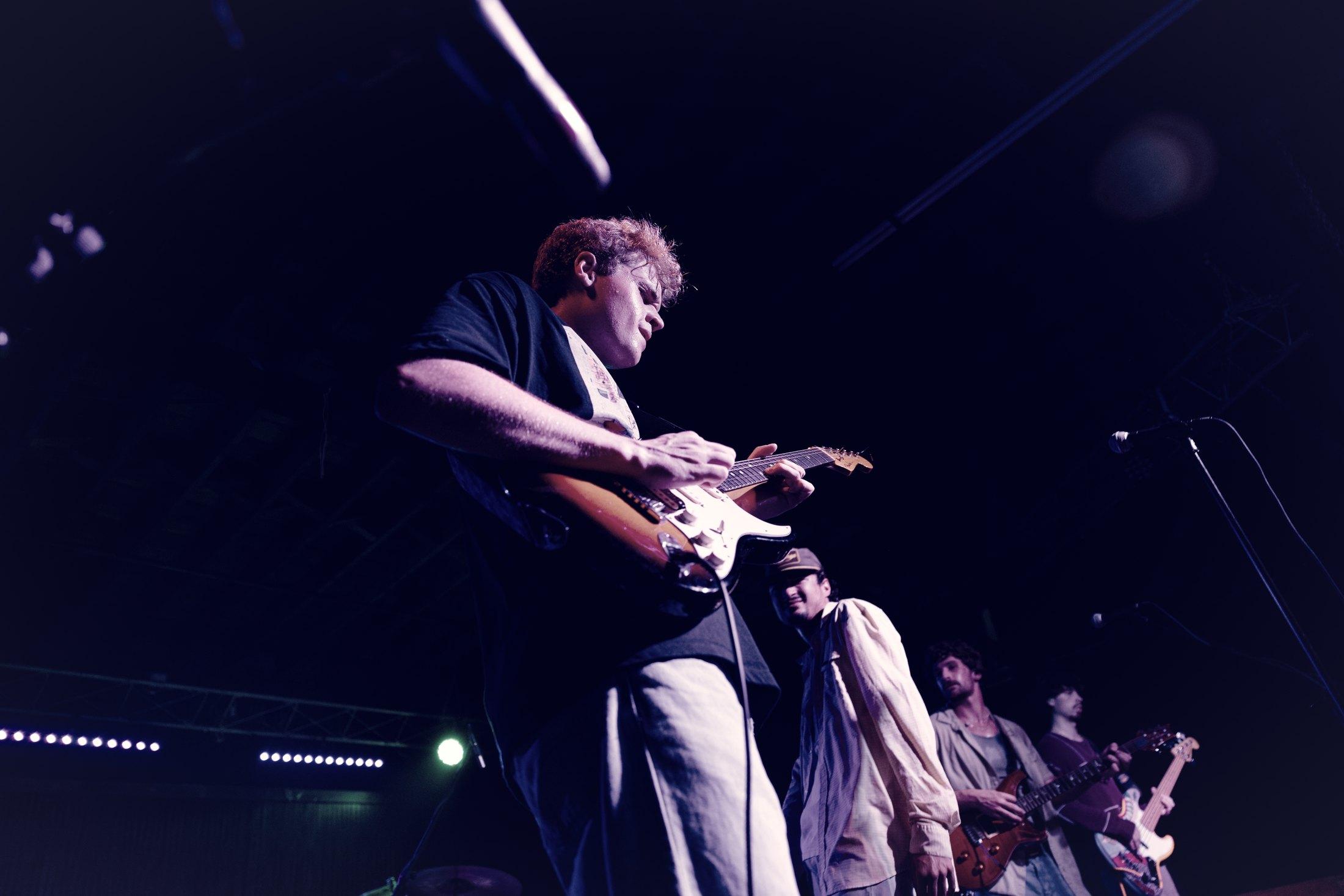A guitarist working at the dark edge of a stage, the band behind him in shadow.